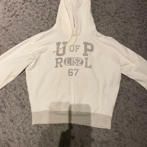 Vit hoodie från Polo Ralph Lauren - Säljer en vit hoodie från Polo Ralph Lauren med tryck på framsidan. Den har en klassisk känguruficka och justerbar huva med snören. Perfekt för en grich stil. Skicket är i 10/10 finns inga defekter, säljer den för 1500 men kan säljas lägre med snabb affär 