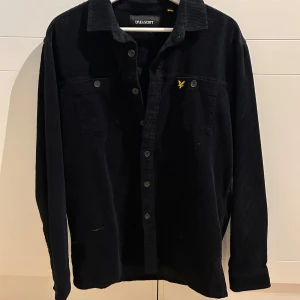 Svart manchester overshirt från Lyle & Scott - Snygg svart overshirt i manchester från Lyle & Scott med klassisk krage och knappar framtill. Den har långa ärmar och en broderad logga på bröstet. Perfekt för lager-på-lager-stil.