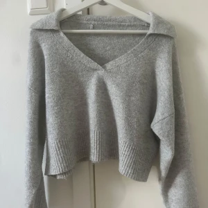 Grå stickad tröja med v-ringning - Säljer en mysig grå stickad tröja med v-ringning och långa ärmar. Tröjan har en croppad design och ribbade detaljer vid ärmslut och nederkant. Perfekt för kyliga dagar!