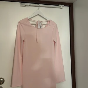 Ljusrosa klänning 'Elina Drapey Dress' - Elegant ljusrosa klänning med långärmad design och en stilren drapering i ryggen. Klänningen har en knytning i nacken och en smickrande passform som passar perfekt för olika tillfällen.     Klänningen har inte kommit till användning , prislapp finns kvar. 