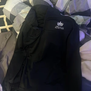 Svart jacka från Active Riding Wear - Snygg svart tröja  från Active Riding Wear med broderad krona och text på bröstet. Perfekt för ridning och andra utomhusaktiviteter. Tröjan har en dragkedja framtill och en hög krage för extra skydd mot väder och vind. Väldigt tunn så perfekt att ha under en jacka