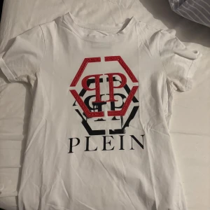 Vit t-shirt från Plein med rött tryck - Snygg vit t-shirt från Plein med ett stort rött och svart tryck på framsidan. T-shirten har korta ärmar och en klassisk rund halsringning. Perfekt för en casual look.