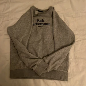 Grå sweatshirt från Peak Performance - Säljer en stilren grå sweatshirt från Peak Performance med tryck på framsidan. Tröjan har långa ärmar och ribbade muddar vid ärmslut och nederkant. Perfekt för en avslappnad stil. Passar runt   Längden 158-165