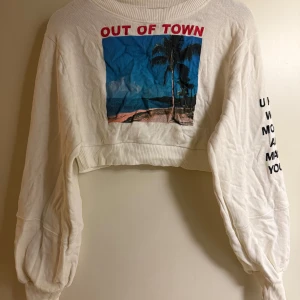 Vit croppad tröja från Bershka - Säljer en vit croppad tröja från Bershka med tryck av en strand och texten 'OUT OF TOWN' i rött. Tröjan har långa ärmar och text på ena ärmen. Perfekt för en avslappnad stil.