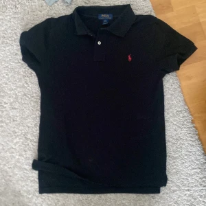 Svart pikétröja från Ralph Lauren - Säljer en klassisk svart pikétröja från Ralph Lauren med ett rött märke tröjan är i väldigt bra skick och storlek L i barnstorlek. Tröjan passar perfekt för dig som är runt 170!