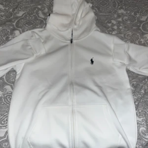 Vit huvtröja från Polo Ralph Lauren - Snygg vit huvtröja från Polo Ralph Lauren med dragkedja och broderad logga på bröstet. Perfekt för en stilren och avslappnad look.