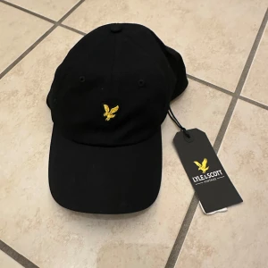 Svart keps från Lyle & Scott - Snygg svart keps från Lyle & Scott med en gul broderad logotyp framtill. Perfekt för en stilren look. Justerbar passform för optimal komfort.