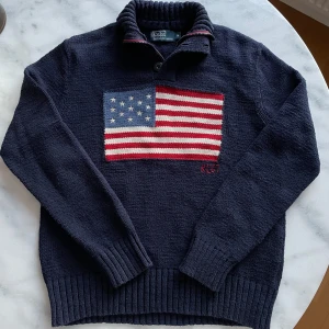 Mörkblå stickad tröja med flagga från Polo Ralph Lauren - Säljer en snygg mörkblå stickad tröja från Polo Ralph Lauren med ett stort amerikanskt flaggmotiv på framsidan. Tröjan har en hög krage med knappar och ribbade muddar vid ärmslut och nederkant. Perfekt för en stilren look!