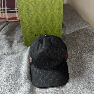 Gucci keps - Snygg svart keps från Gucci med det ikoniska GG-mönstret. Kepsen har gröna och röda ränder på sidan och justerbar rem baktill. Perfekt för en stilren look.