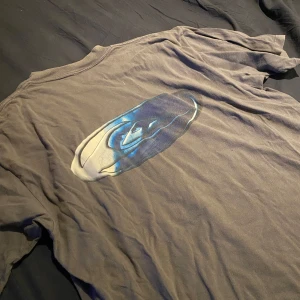 Grå t-shirt från Quiksilver - Säljer en grå t-shirt från Quiksilver med ett coolt tryck på baksidan. T-shirten har korta ärmar och en liten logga på framsidan. Perfekt för en avslappnad stil. Lite lös i kragen.