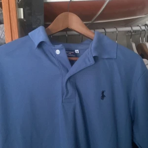 Blå pikétröja från Ralph Lauren - Säljer en klassisk blå pikétröja från Ralph Lauren. Tröjan är tillverkad i 100% bomull och har en broderad logotyp på bröstet. Perfekt för en stilren och avslappnad look.