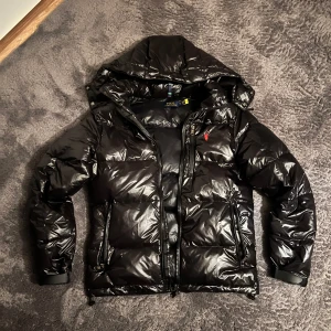 Polo Ralph Lauren jacka - Snygg svart pufferjacka från Polo Ralph Lauren med glansig yta och huva. Jackan har dragkedja och två fickor framtill. Perfekt för kyliga dagar.