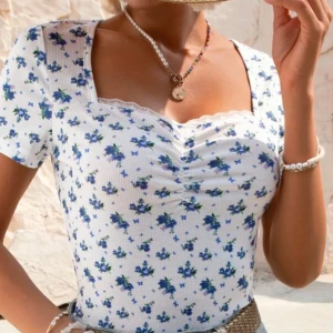 Vit blommig topp - Söt vit topp med blått blommönster och spetsdetaljer vid halsringningen. Toppen har korta ärmar och en ribbad struktur som ger en fin passform. Perfekt för en somrig look!