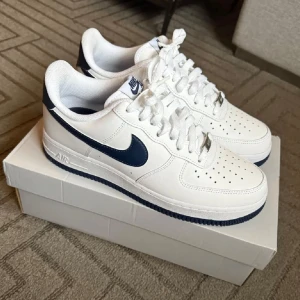 Nike Air Force 1 '07 i vitt och midnattsblått - Snygga Nike Air Force 1 '07 sneakers i vitt med midnattsblå detaljer. Skorna har en klassisk design med snörning och perforerade detaljer på tån för extra ventilation. Perfekta för en stilren look.