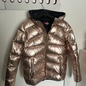 Roséguldig pufferjacka från Colmar - Snygg roséguldig pufferjacka från Colmar med dragkedja och huva. Jackan har en glansig finish och är perfekt för kyligare dagar. Insidan är mörkblå vilket ger en fin kontrast. Den är nästintill oanvänd och köptes i en Colmarbutik i Italien.