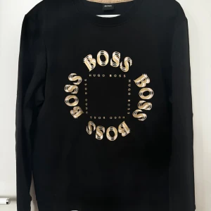 Hugo Boss tröja strl M - Snygg svart tröja från Hugo Boss med ett unikt cirkulärt mönster av logotypen på framsidan. Tröjan har långa ärmar och en klassisk rund halsringning. Perfekt för en stilren look.