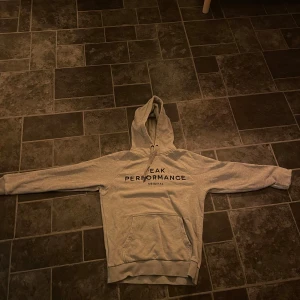Grå hoodie från Peak Performance - Säljer en grå hoodie från Peak Performance med text på framsidan. Den har en klassisk känguruficka och en bekväm huva. Perfekt för en avslappnad stil. Jätte tunt tyg (trasig) 99 armbågarna
