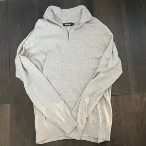Halv zip - Säljer en stilren grå tröja från Dressmann med dragkedja vid halsen. Tröjan har långa ärmar och ribbade muddar vid ärmslut och nederkant. Perfekt för en avslappnad och bekväm stil. Den är i storlek M och är knappt använd