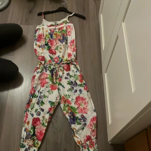 Blommig jumpsuit från Kids by Lindex - Säljer en färgglad blommig jumpsuit från Kids by Lindex. Den har ett vackert blommönster i rosa, grönt och blått på en vit bakgrund. Jumpsuiten har smala axelband och ett knytband i midjan för justerbar passform. Perfekt för sommarens varma dagar!