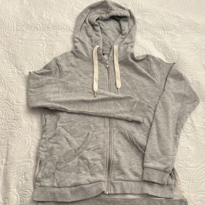 Grå hoodie från Lindex - Säljer en grå hoodie från Lindex i storlek M. Den har en dragkedja framtill och justerbara snören i huvan samt små slits på varsin sida. Tillverkad i mjuk bomullsmix för extra komfort.