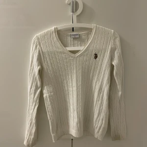 Vit stickad tröja från U.S. Polo Assn. - Säljer en vit stickad tröja från U.S. Polo Assn. med klassiskt kabelstickat mönster och v-ringning. Tröjan har en broderad logotyp på bröstet och långa ärmar. Perfekt för en elegant och avslappnad look. Är lite fläckig vid ärmarna men annars helt stilren