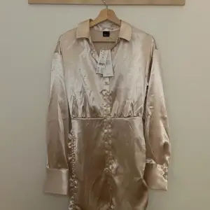 Elegant champagnefärgad skjortklänning i satin från Gina Tricot. Klänningen har långa ärmar och knappar framtill, vilket ger en stilren look. Perfekt för en chic och sofistikerad stil.