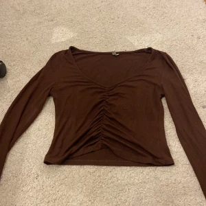 Brun långärmad topp från NLY Trend - Säljer en brun långärmad topp från NLY Trend med v-ringning och rynkdetalj framtill. Perfekt för en stilren look.