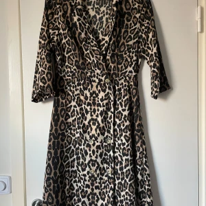 Leopardmönstrad klänning från ONLY - Snygg leopardmönstrad klänning från ONLY med trekvartsärmar och omlott-design. Perfekt för att sticka ut med stil. Klänningen har en v-ringad hals och knappar framtill.