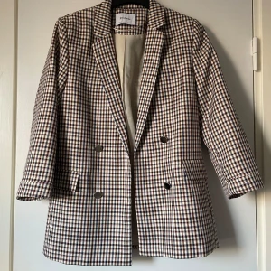 Rutig kavaj från Stradivarius - Snygg rutig kavaj från Stradivarius i beige, blå och brun. Kavajen har en klassisk design med dubbelknäppning och långa ärmar. Perfekt för en stilren look.