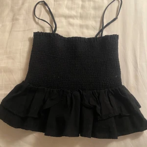 Svart axelbandslös topp - Snygg svart axelbandslös topp med smockdetaljer och volangkant. Perfekt för en somrig look med tunna axelband.