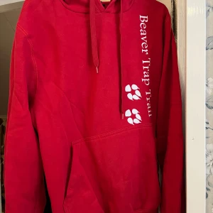 Röd hoodie med tryck - Säljer en röd hoodie med texten 'Beaver Trap Trail' och ett tryck av tassavtryck på framsidan. Baksidan har ett stort runt tryck med naturmotiv och text. Perfekt för en avslappnad stil.