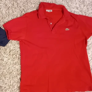 Röd pikétröja från Lacoste - Snygg röd pikétröja från Lacoste med klassisk krage och knappdetalj. Den har det ikoniska broderade krokodilemblem på bröstet. Perfekt för en stilren och avslappnad look.
