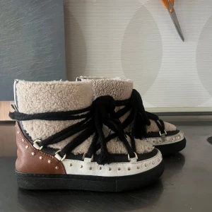 Inuikii skor curly rock - Nypris runt 4000 kr, endast använda max 10 gånger. Dem var inga fel eller märken! Storlek 36 men passar upp till 38 Som nya Snygga och varma boots med en unik design. De har en beige och brun färgkombination med svarta snören. Skorna är dekorerade med nitar och har en mysig fodring som ger extra värme. Perfekta för kyligare dagar.