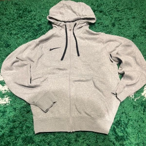 Grå Zip hoodie från Nike - Säljer en stilren grå hoodie från Nike med dragkedja och svart logga på bröstet. Den har en justerbar huva med svarta snören och långa ärmar. 