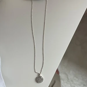 Silverfärgat halsband med glittrig berlock - Elegant silverfärgat halsband med en rund, glittrig berlock. Perfekt för att ge en touch av glamour till din outfit. Kedjan är tunn och smidig. 