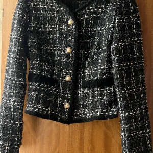 Svartvit rutig kavaj med knappar - Snygg svartvit rutig kavaj med dekorativa knappar. Den har en klassisk design med långa ärmar och två fickor framtill. Perfekt för en stilren look.