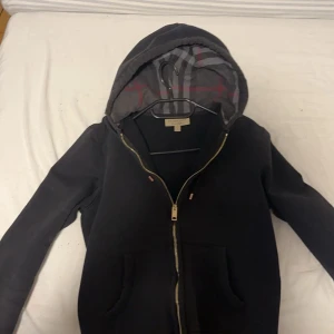 Svart burberry hoodie - Säljer en stilren svart huvtröja från Burberry med dragkedja och klassiskt rutigt mönster i huvan. Tröjan har långa ärmar och en diskret broderad logga på bröstet. Perfekt för en casual look. Tar även byten mot den