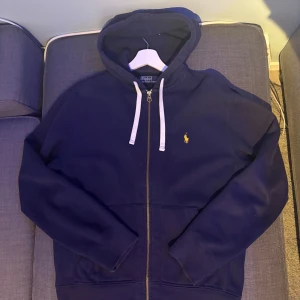 Ralph Lauren Zip Hoodie - Säljer en snygg mörkblå zip hoodie från Ralph Lauren. Den har en broderad gul logga på bröstet samt vita snören. Fint skick. Storlek M!