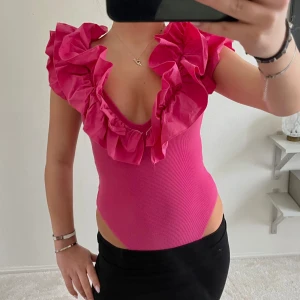 Rosa volangbody Zara - Säljer en snygg rosa bodysuit med volangdetaljer och djup v-ringning. Perfekt för att sticka ut med sin livfulla färg och unika design. Passar bra till både kjol och byxor.
