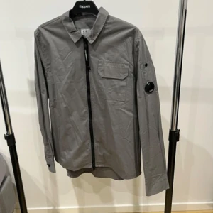 Grå CP company overshirt - Helt ny och oanvänd CP company overshirt, 10/10 skick. Den har inga skador eller liknande. Den är lite skrynklig pga den legat i garderoben ett tag. Men de går enkelt att stryka den med låg temperatur. Riktigt bekväm och fin, rekommenderar starkt!