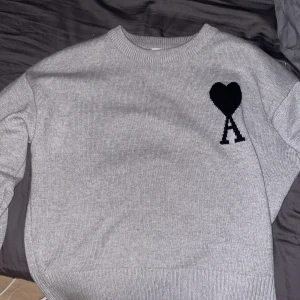 Grå stickad tröja från Ami - Säljer en stilren grå stickad tröja från Ami med ett svart hjärta och bokstaven 'A' på bröstet. Tröjan har en rund halsringning och långa ärmar, perfekt för kyligare dagar. Ett måste för garderoben! 🖤