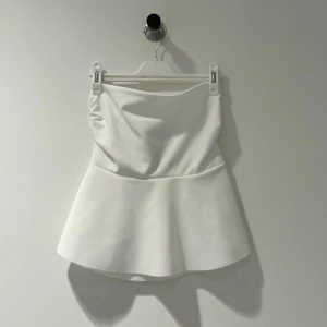 Vit axelbandslös topp - Snygg vit axelbandslös topp med en peplumdetalj som ger en elegant siluett. Perfekt för att klä upp en outfit med sin stilrena design.