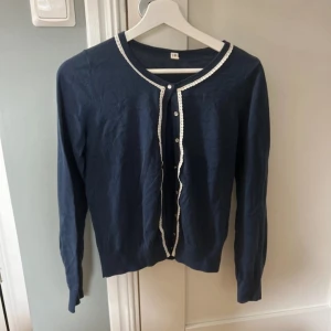 Mörkblå kofta från Espirit - Säljer en elegant mörkblå kofta med vita spetsdetaljer längs knäppningen och halsringningen. Koftan har långa ärmar och knappar framtill. Perfekt för en stilren look.