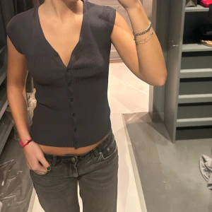 Mörkgrå ribbad topp med knappar - Snygg mörkgrå ribbad topp med knappar framtill. Toppen har en v-ringad design och är ärmlös, vilket ger en stilren look. Perfekt för en avslappnad stil.