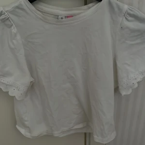 Vit topp från Vero Moda - Säljer en söt vit topp från Vero Moda med korta puffärmar och spetsdetaljer vid ärmsluten. Perfekt för en somrig look. Toppen har en rund halsringning och är gjord i ett mjukt material.