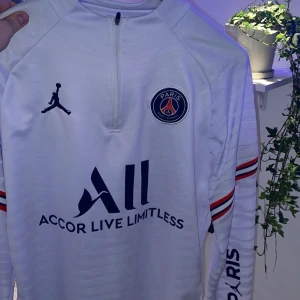 Vit långärmad PSG-tröja - Säljer en vit långärmad PSG-tröja med Jordan-logga och Paris Saint-Germain-emblem på bröstet. Tröjan har texten 'Accor Live Limitless' och 'Visit Rwanda'. Den har även ränder i rött och blått på ärmarna. Perfekt för fotbollsfans!