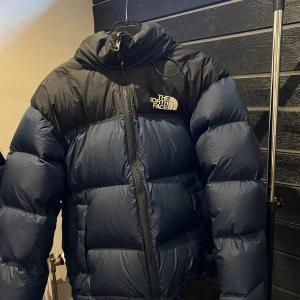Svart och blå dunjacka från The North Face - Säljer en snygg och varm dunjacka från The North Face i svart och blått. Jackan har en hög krage och dragkedja framtill. Perfekt för kalla dagar och håller dig varm och bekväm. 🧥
