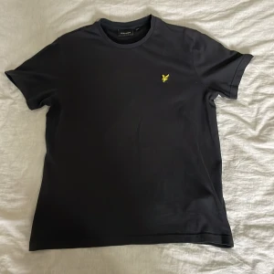 Svart t-shirt från Lyle & Scott - Säljer en stilren svart t-shirt från Lyle & Scott med en gul broderad logga på bröstet. Perfekt för en avslappnad look. T-shirten är kortärmad och gjord i mjukt material. Storlek M, nypris 399kr, djur och rökfritt hem