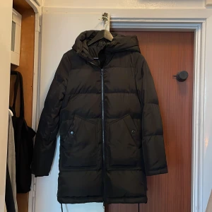 Svart dunjacka från Vero Moda - Säljer en svart dunjacka från Vero Moda med huva och dragkedja framtill. Jackan har långa ärmar och två stora fickor med knappar. Perfekt för kalla dagar.  Strl S/M