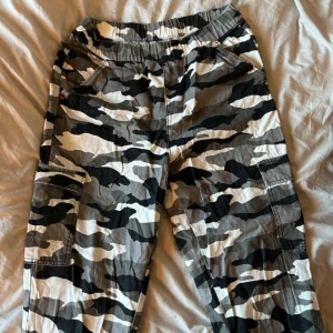 Camouflage cargobyxor - Snygga cargobyxor i ett klassiskt grått och svart camouflagemönster. Byxorna har elastisk midja för extra komfort och praktiska fickor på sidorna. Perfekta för en avslappnad stil.